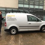 Oliver Broadbent stands next to a van that has the caption on it Creativty with Plymouth Univesity. Oliver is pointing at the word creativity. He was invited to the university to talk about teaching conceptual design