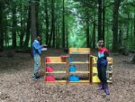Two participants at the regenerative design lab stand in front of three bookcases situated in a forest clearing. The books on the left bookcase are red, in the middle are blue and on the bookcase on the right they are yellow. The different colours represent H1, H2 and H3 respectively in the Three Horizons model