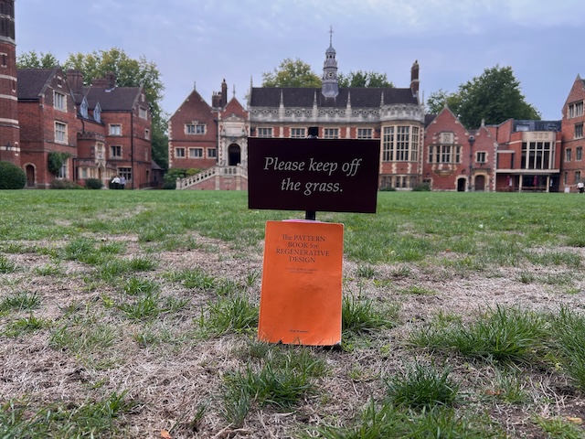 The Pattern Book for Regenerative Design is propped against a sign saying keep off the grass. In the backdrop is the quad of a Cambridge college