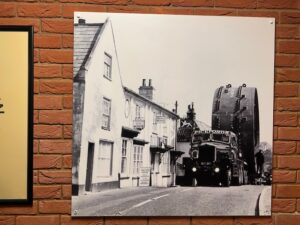 Black and white photo showing a large cycling on the back of a lorry being towed through a village. The cylinder is much larger than the surrounding houses.