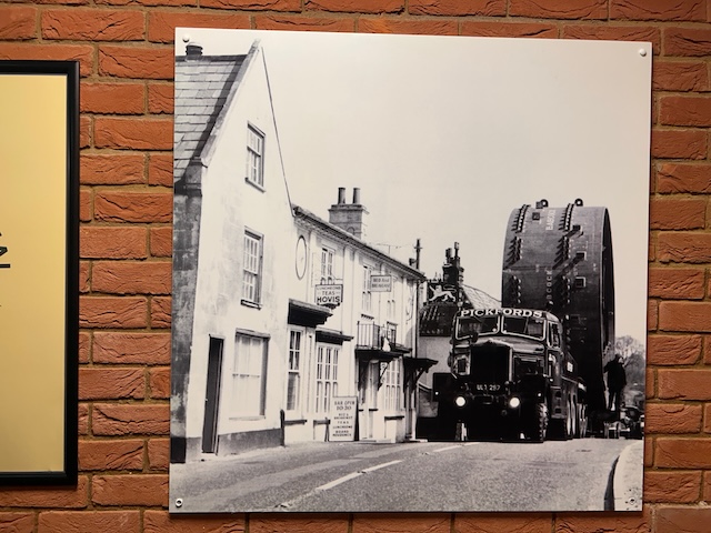 Black and white photo showing a large cycling on the back of a lorry being towed through a village. The cylinder is much larger than the surrounding houses.