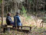Two people sit on a bench near a pond in a wood, to emphasise the importance of connecting with ecosystems